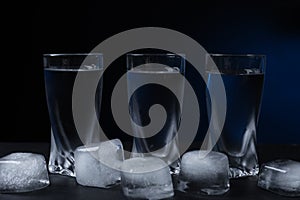 Shot glasses of vodka with ice cubes on black table against dark background