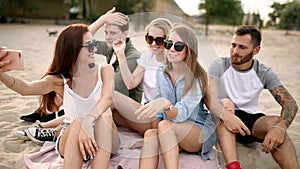 Shot of a group of young friends taking a selfie on the beach. Men and women taking photos sitting on a sand on warm