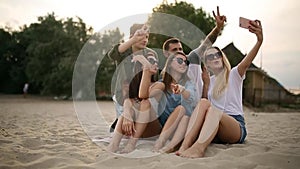 Shot of a group of young friends taking a selfie on the beach. Men and women taking photos sitting on a sand on warm