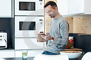 Handsome mature business man eating take away noodles while using smartphone in the kitchen at home