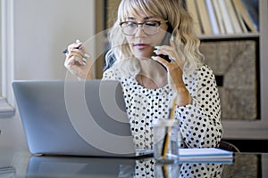 Shot happy businesswoman sitting at desk behind her laptop and talking with somebody on her mobile phone while working from home.
