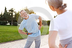 Shot of happy smiling mature family man and woman in sportswear stretching arms while warming up together outdoors in park on sunn