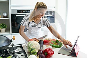 Healthy young woman cutting fresh vegetables and using digital tablet to recipes in the kitchen at home.