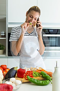 Healthy young woman eating a piece of fresh vegetables and talking with her mobile phone in the kitchen at home.