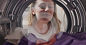 A shot from inside the drum. A woman holds a laundry capsule which she puts into colorful clothes lying around, a girl