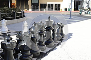 A shot of a large black and white chess set on the sidewalk surrounded by lush green plants, bare winter trees and shops