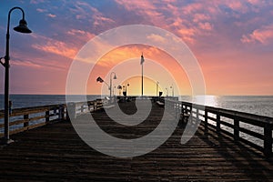 A shot of a long brown wooden pier at sunset with vast blue ocean water and American flags on curved light posts