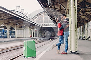 Shot of lovely boyfriend and girlfriend stand closely, embrace and kiss, wait for transport on railway station, being passengers,