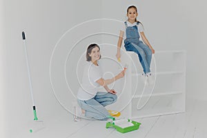 Shot of lovely mother and small hard working daughter pose in empty room, paint furniture in white color, use paintbrushes, happy