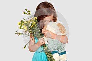 Shot of lovely small female child plays with her favourite toy, holds beautiful flowers, enjoys recieveing present, dressed in