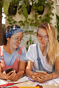 Shot of multiethnic females share multimedia files via bluetooth, hold modern cellulars, sit at desktop, collaborate