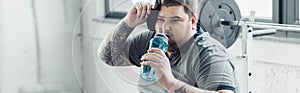 Shot of overweight tattooed man drinking water from sport bottle and wiping face with towel at gym
