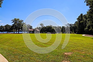 A shot of a park by the lake with vast green grass, lush green trees, people relaxing on the banks of the lake, blue sky and cloud