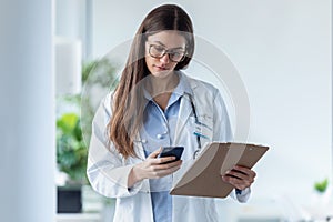 Pretty female doctor using her mobile phone while reviewing medical reports in medical consultation