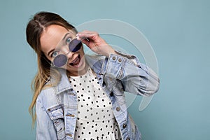 Shot of pretty positive joyful young blonde woman wearing blue jean jacket and stylish sunglasses isolated over blue
