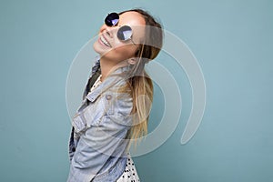 Shot of pretty positive smiling young blonde woman wearing blue jean jacket and stylish sunglasses isolated over blue