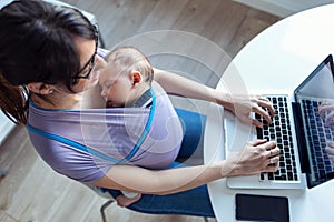 Pretty young mother with her baby in sling working with laptop at home