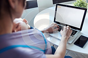 Pretty young mother with her baby in sling working with laptop at home