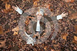 Pretty young woman with her dog looking to the sky with arms raised as leaves fall from the trees in the park in autumn.