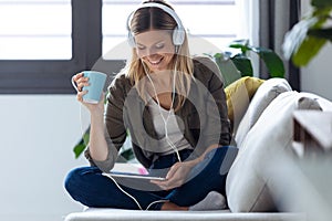 Pretty young woman listening to music with her digital tablet while drinking cup of coffee on sofa at home