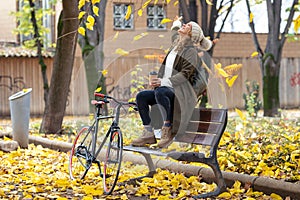 Pretty young woman looking to the sky with arms raised as leaves fall from the trees sitting on bench in the park in autumn.