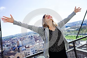 Pretty young woman with raised arms to the sky enjoying the time at modern rooftop of high-rise building.