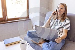 Shot of pretty young woman using her mobile phone while working with laptop sitting on comfortable couch at home