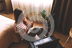 Shot of pretty young woman using her mobile phone while working with laptop sitting on the floor at home.