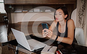 Shot of pretty young woman using her mobile phone while working with laptop sitting at home