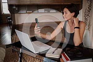 Shot of pretty young woman using her mobile phone while working with laptop sitting at home