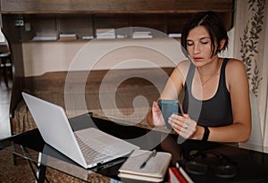 Shot of pretty young woman using her mobile phone while working with laptop sitting at home