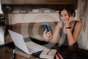 Shot of pretty young woman using her mobile phone while working with laptop sitting at home.