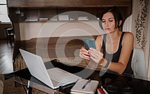 Shot of pretty young woman using her mobile phone while working with laptop sitting at home