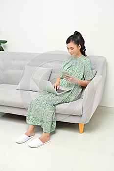 Shot of pretty young woman using her mobile phone while working with laptop sitting on sofa at home