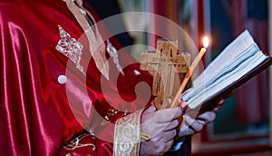 Shot of a priest in a red cassock reading a prayer with a wooden cross, and candle in hands