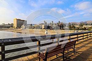 A shot of seagulls standing on the railing of a long brown wooden pier with ocean water rolling into the beach with hotels