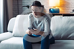 Serious young woman using her mobile phone while sitting on sofa in the living room at home