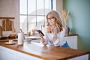 Smiling woman text messaging and drinking morning coffee while standing in the kitchen at home