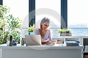 Smiling young business woman using her mobile phone while working with computer in the office at home