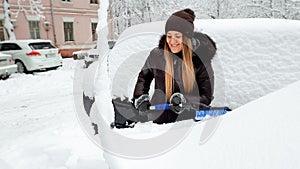 Shot of smiling young woman in coat jeans and hat trying to clean up snow covered white car by blue brush after a