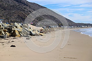 A shot of the smooth sand at the beach with large rocks along the sand near a large mountain range and RVs parked