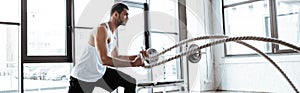 Shot of strong sportsman exercising with battle ropes in modern gym