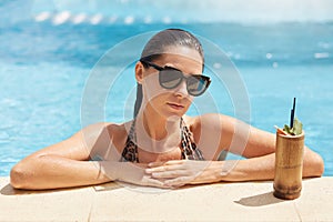 Shot of stunning beautiful young woman enjoying sunbathing in pool with fresh fruit cocktail, cheerful attractive woman having