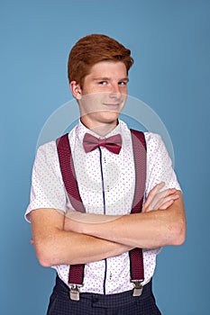 Shot of a  stylish red-haired young smiling boy teenager wearing  white shirt with a red bow tie and suspenders  with arms crossed