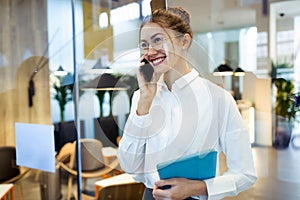 Stylish young businesswoman talking with her mobile phone while holding her digital tablet standing in the hotel lobby