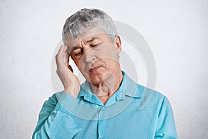 Shot of tired elderly male pensioner, keeps eyes closed, hand on temple, wears formal blue shirt, has headache, poses against whit