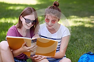 Shot of two cheerful friends have joyful looks into book, read something funny, wear sunglasses, pose against green grass backgrou