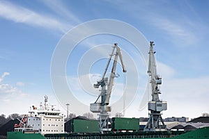 Shot of two old, rusty, grey port cranes with big hooks, lifting cargo in ship on clear blue sky background.