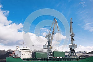 Shot of two old, rusty, grey port cranes with big hooks, lifting cargo in ship on clear blue sky background.