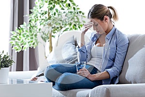 Unhealthy young woman with headache holding glass of water while sitting on sofa at home.